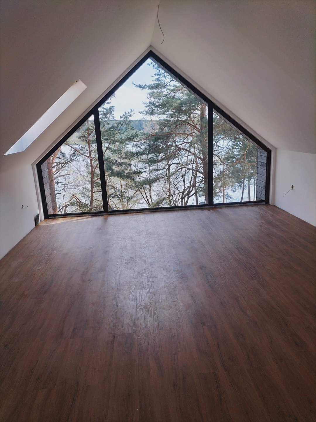 Poddasze z oknem w kształcie trójkąta, z widokiem na jezioro i drzewa. Na podłodze ułożone ciemnobrązowe panele.