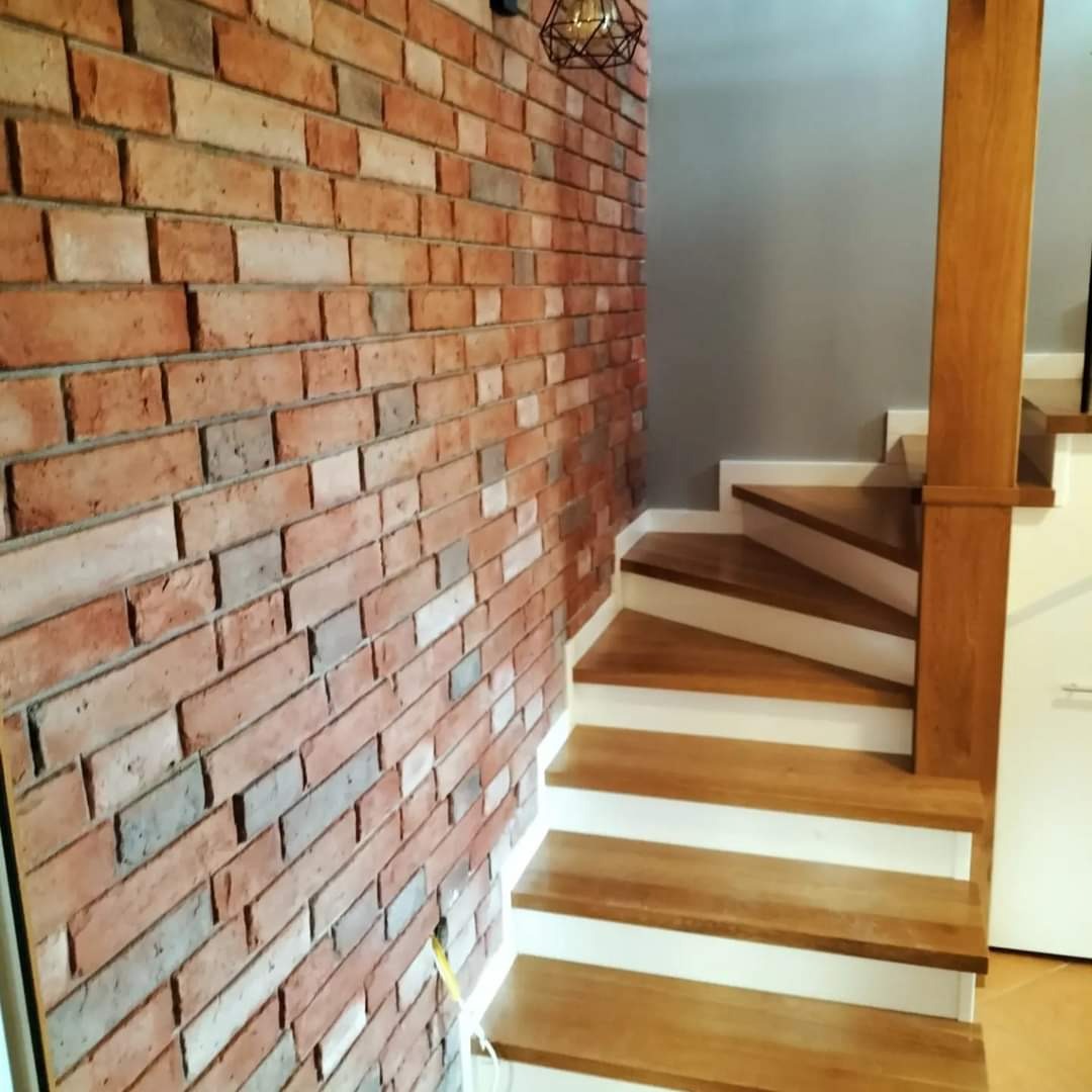 Ściana z czerwonej cegły dekoracyjnej sąsiaduje z drewnianymi schodami z białymi podstopnicami, oświetlonymi przez designerską lampę wiszącą.