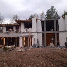 GRINKOR Sp. zo.o - Dwukondygnacyjny budynek w stanie surowym otwartym z białych bloczków, widoczne drewniane szalunki, podpory i sterta desek na ziemi, teren budowy z liśćmi.