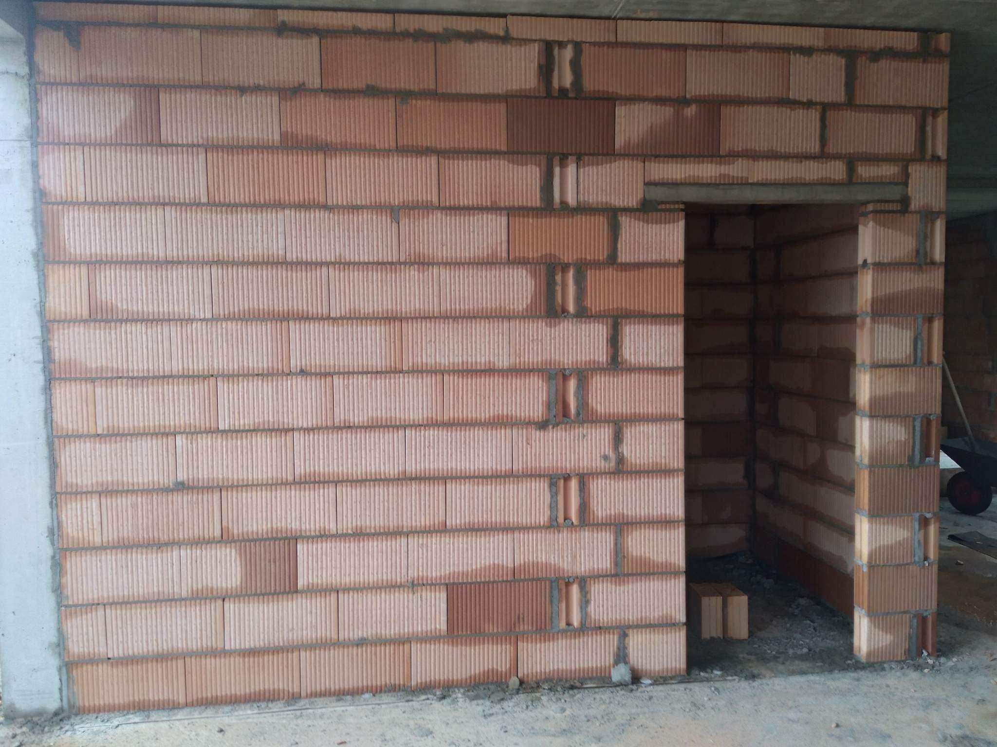 Ściana z czerwonych bloczków ceramicznych Porotherm w trakcie wznoszenia, widoczny otwór drzwiowy i fragment betonowego słupa na krawędzi ściany.