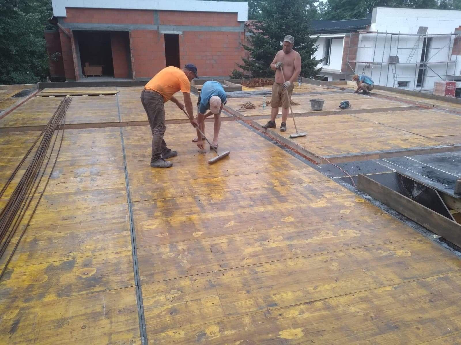 Trzech robotników zamiata deskowanie szalunkowe pod wylewkę stropu na budowie domu murowanego, widoczne zbrojenie i fragmenty ścian.