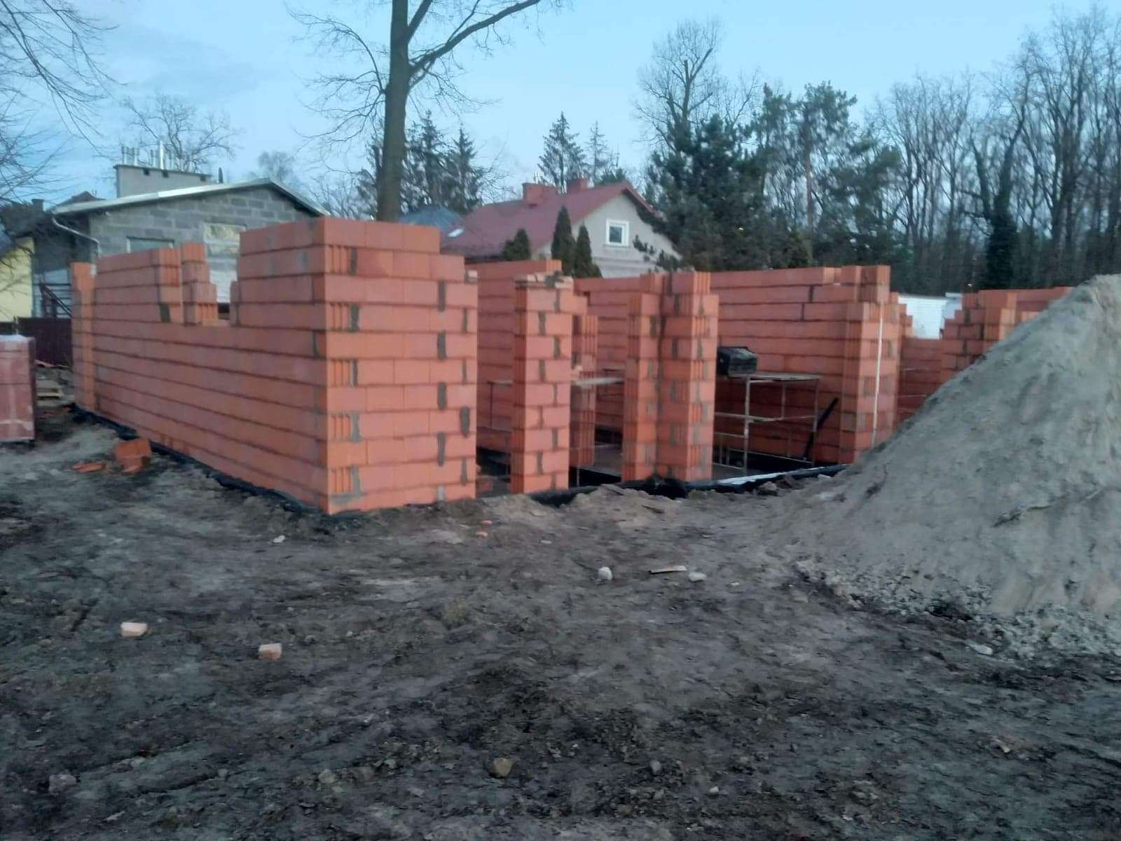 Wznoszone ściany domu z czerwonej cegły, widoczne rusztowania i hałda piasku na placu budowy w pochmurny dzień.