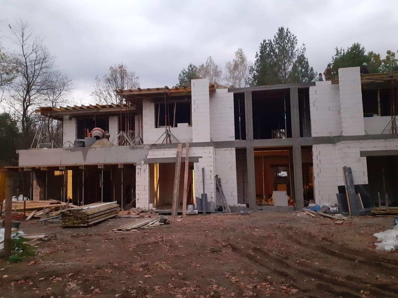 Dwukondygnacyjny budynek w stanie surowym otwartym z białych bloczków, widoczne drewniane szalunki, podpory i sterta desek na ziemi, teren budowy z liśćmi.