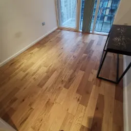 Świeżo ułożona, jasna podłoga z paneli w nowoczesnym mieszkaniu z dużym oknem i fragmentem czarnego stolika kawowego