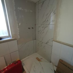 Krzysztof Zarski - Wnętrze łazienki w trakcie remontu: częściowo ułożone płytki imitujące marmur na ścianach, surowy tynk przy oknie, czerwona skrzynka narzędziowa i karton z płytkami na podłodze.