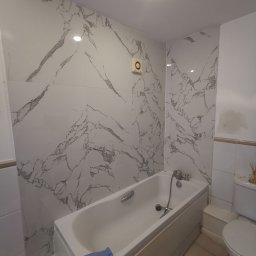 Krzysztof Zarski - Biała wanna obudowana płytkami, ściany wyłożone płytkami imitującymi marmur, widoczny fragment toalety, niebieska ściereczka leżąca na obudowie wanny.