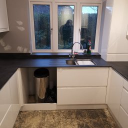 Krzysztof Zarski - Fragment nowej, białej zabudowy kuchennej z czarnym blatem, widoczny zlew, bateria i okno z witrażami, pod zlewem kosz na śmieci; podłoga w trakcie prac wykończeniowych.