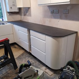 Krzysztof Zarski - Częściowo zamontowane białe szafki kuchenne z czarnym blatem w trakcie remontu kuchni w Braniewie, widoczne narzędzia i nieukończone ściany.