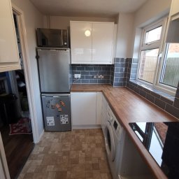 Krzysztof Zarski - Niewielka kuchnia z białymi szafkami, drewnianym blatem, szarą glazurą i pralką pod blatem, widok na lodówkę z magnesami