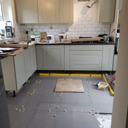 Krzysztof Zarski - Montaż szarych płytek podłogowych w kuchni z użyciem poziomicy i klinów dystansowych, widoczne szafki kuchenne w odcieniach szarości i białe płytki na ścianie w trakcie układania.