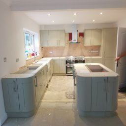 Krzysztof Zarski - Wnętrze kuchni w trakcie montażu: szare szafki z zaokrąglonymi rogami, wyspa kuchenna bez blatu, widoczne przygotowania do położenia płytek nad blatem, surowa podłoga.