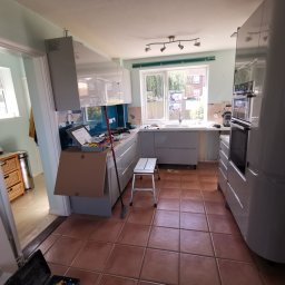 Krzysztof Zarski - Kuchnia w trakcie montażu: szare szafki kuchenne z połyskiem, widoczne narzędzia, drabinka i kartonowe osłony, płytki częściowo zdjęte ze ściany nad blatem, terakota na podłodze.