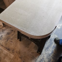 Krzysztof Zarski - Fragment blatu roboczego z zaokrąglonym brzegiem, wspartego na kozłach stolarskich, w warsztacie z widocznymi wiórami na podłodze.