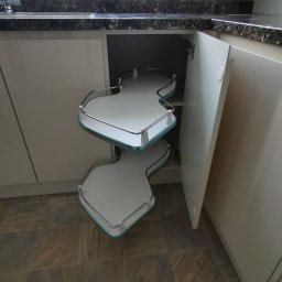 Krzysztof Zarski - Wysuwana półka narożna w szafce kuchennej z otwartymi drzwiczkami, widoczne dwie białe półki z chromowanymi relingami i jasnoniebieskim obramowaniem.