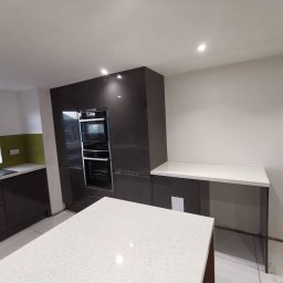 Krzysztof Zarski - Nowoczesna kuchnia z ciemnymi, lakierowanymi frontami, wbudowanym piekarnikiem i wyspą z białym blatem w trakcie montażu.