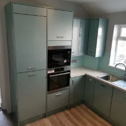 Nowoczesna kuchnia w odcieniach pastelowej zieleni z zabudowanymi sprzętami AGD, wykończona panelami podłogowymi o wzorze drewna.