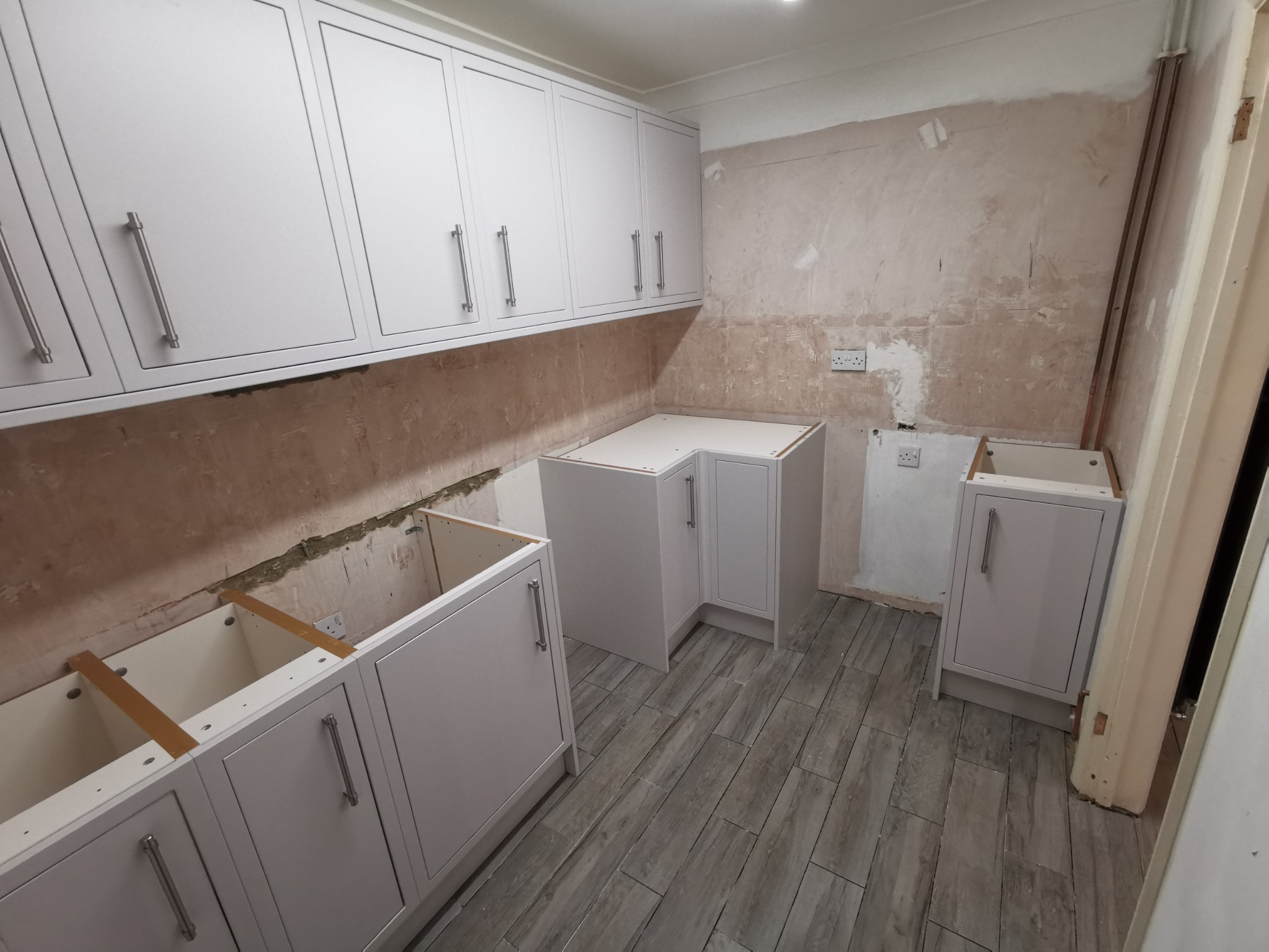 Montaż jasnych szafek kuchennych na tle surowych ścian, widoczne drewniane elementy mocujące i szare panele podłogowe.