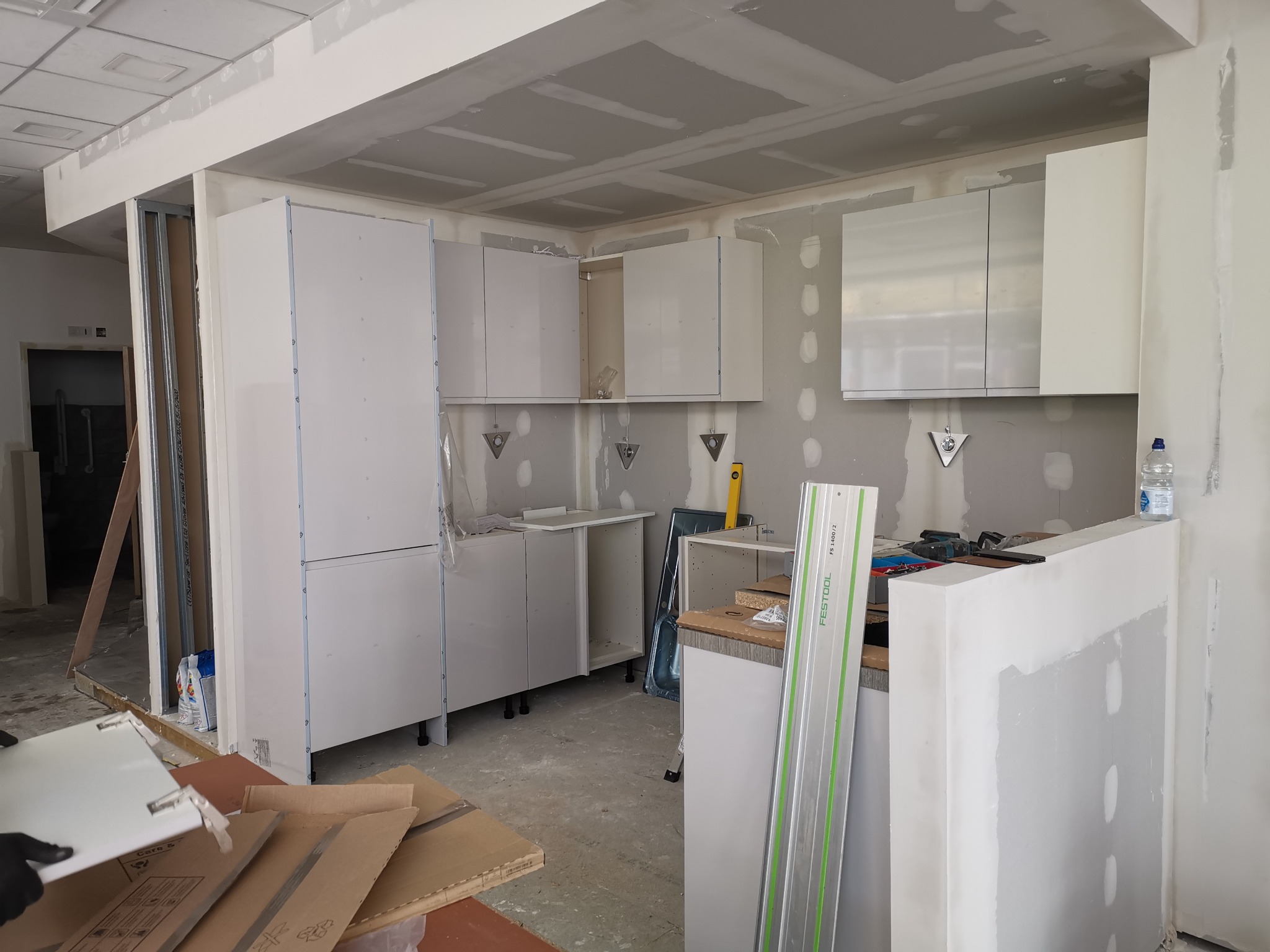 Montaż białych szafek kuchennych w surowym wnętrzu, widoczne narzędzia i materiały budowlane, ściany w trakcie wykończenia, fragment podłogi z kartonami.