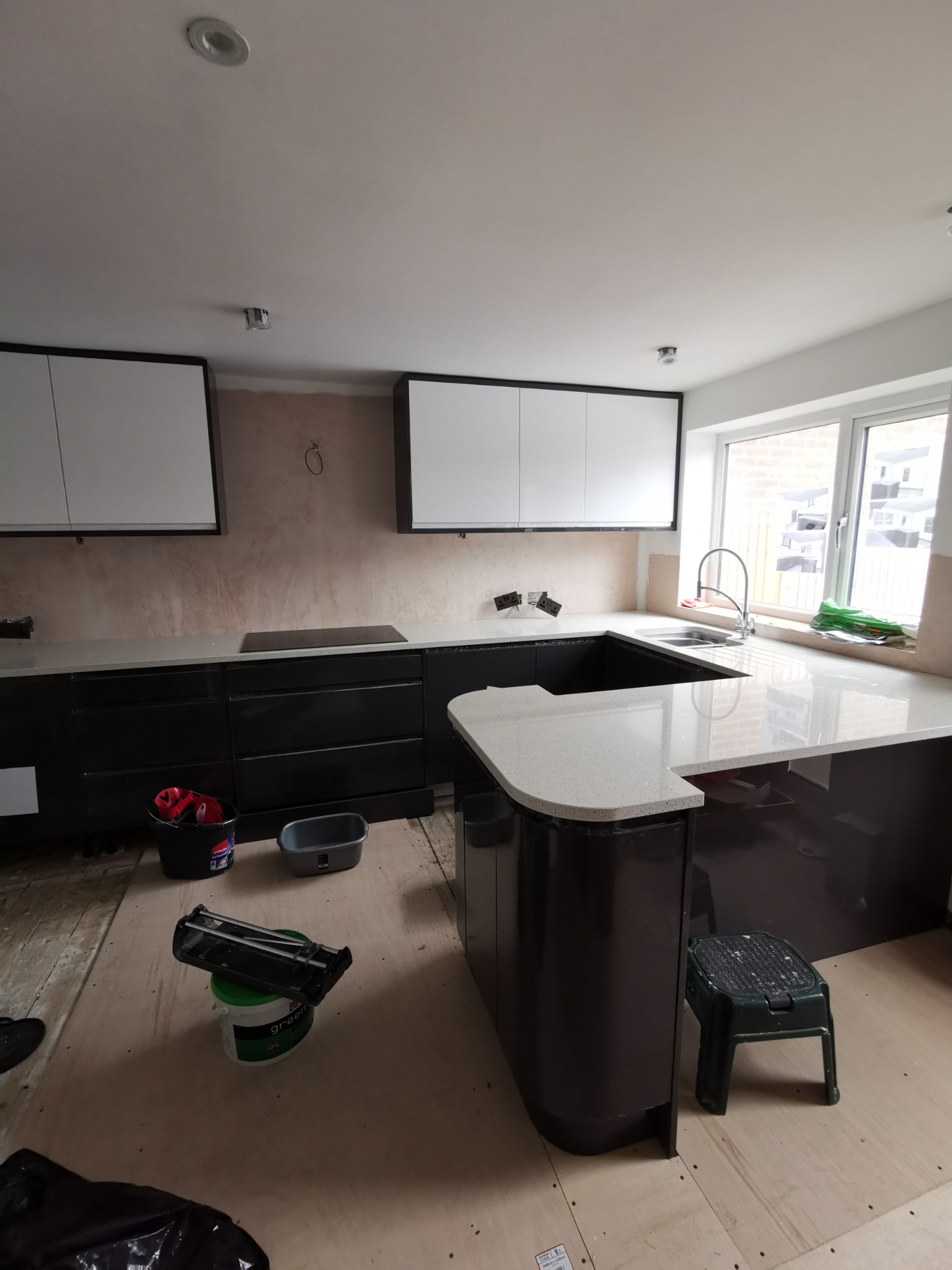 Częściowo zamontowana kuchnia z czarnymi szafkami dolnymi i białymi górnymi, wyspą z blatem w kolorze jasnego lastryko, widoczne narzędzia i materiały budowlane na podłodze.