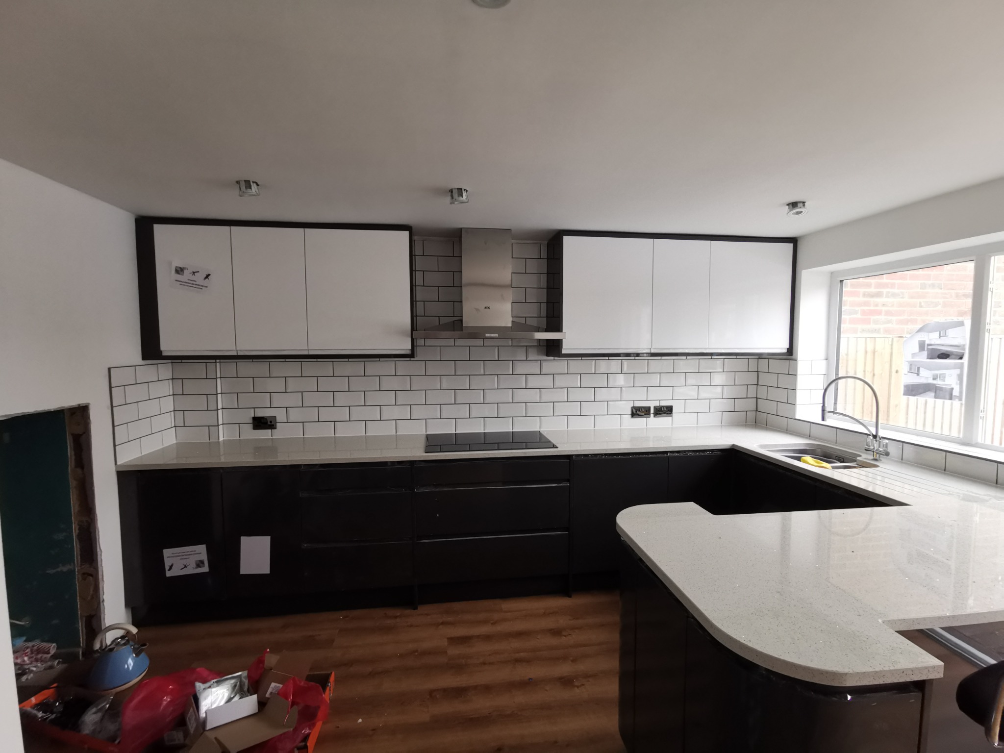 Częściowo ukończona kuchnia z czarnymi szafkami dolnymi i białymi górnymi, białą mozaiką na ścianie, stalowym okapem i blatem w jasnym odcieniu szarości; widoczne elementy instalacji elektrycznej...