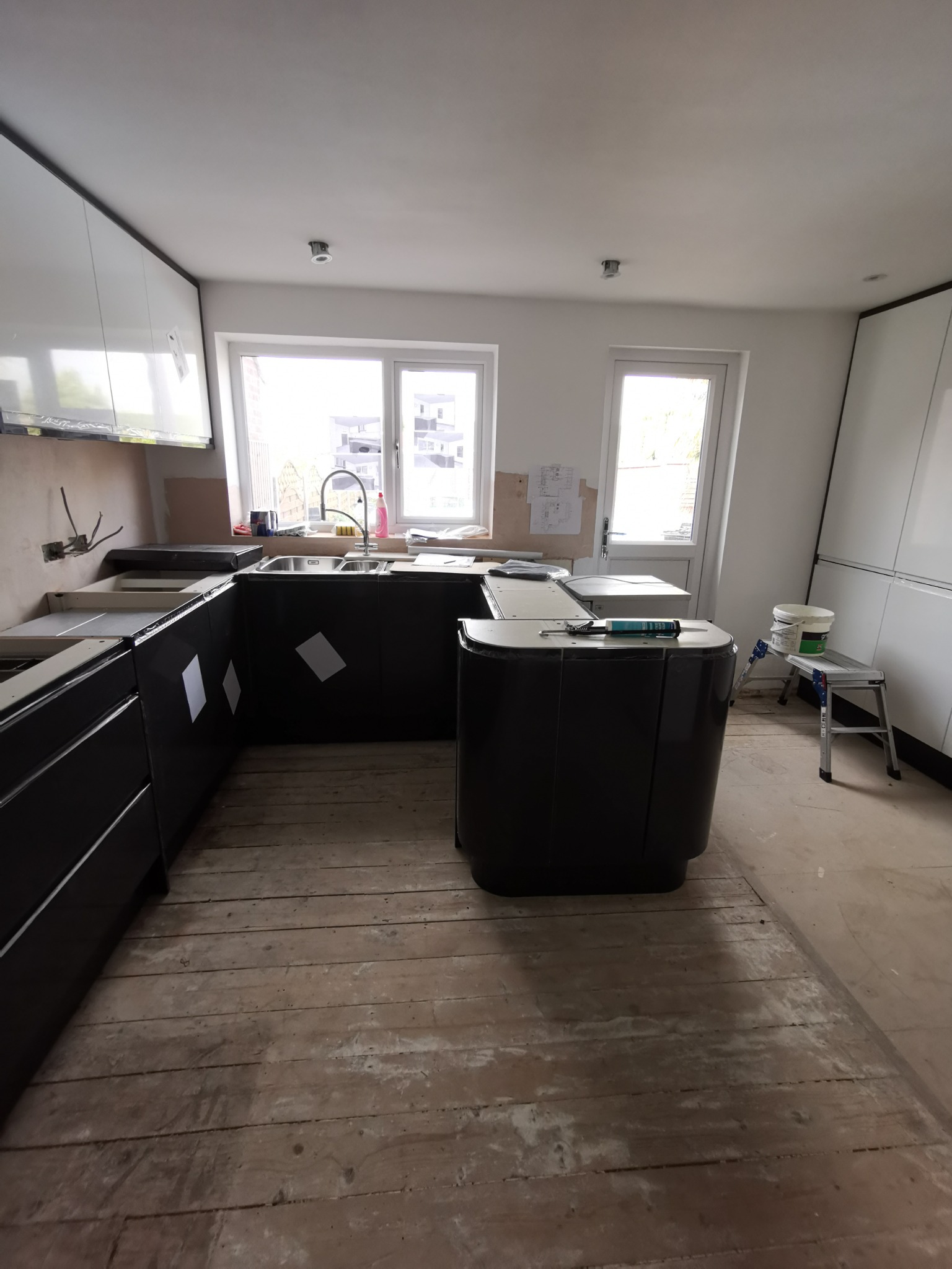 Częściowo zamontowana, nowoczesna kuchnia z czarnymi szafkami i białymi górnymi szafkami, widoczne przygotowania do montażu blatu, surowe drewniane podłogi i drabina z farbą w tle.