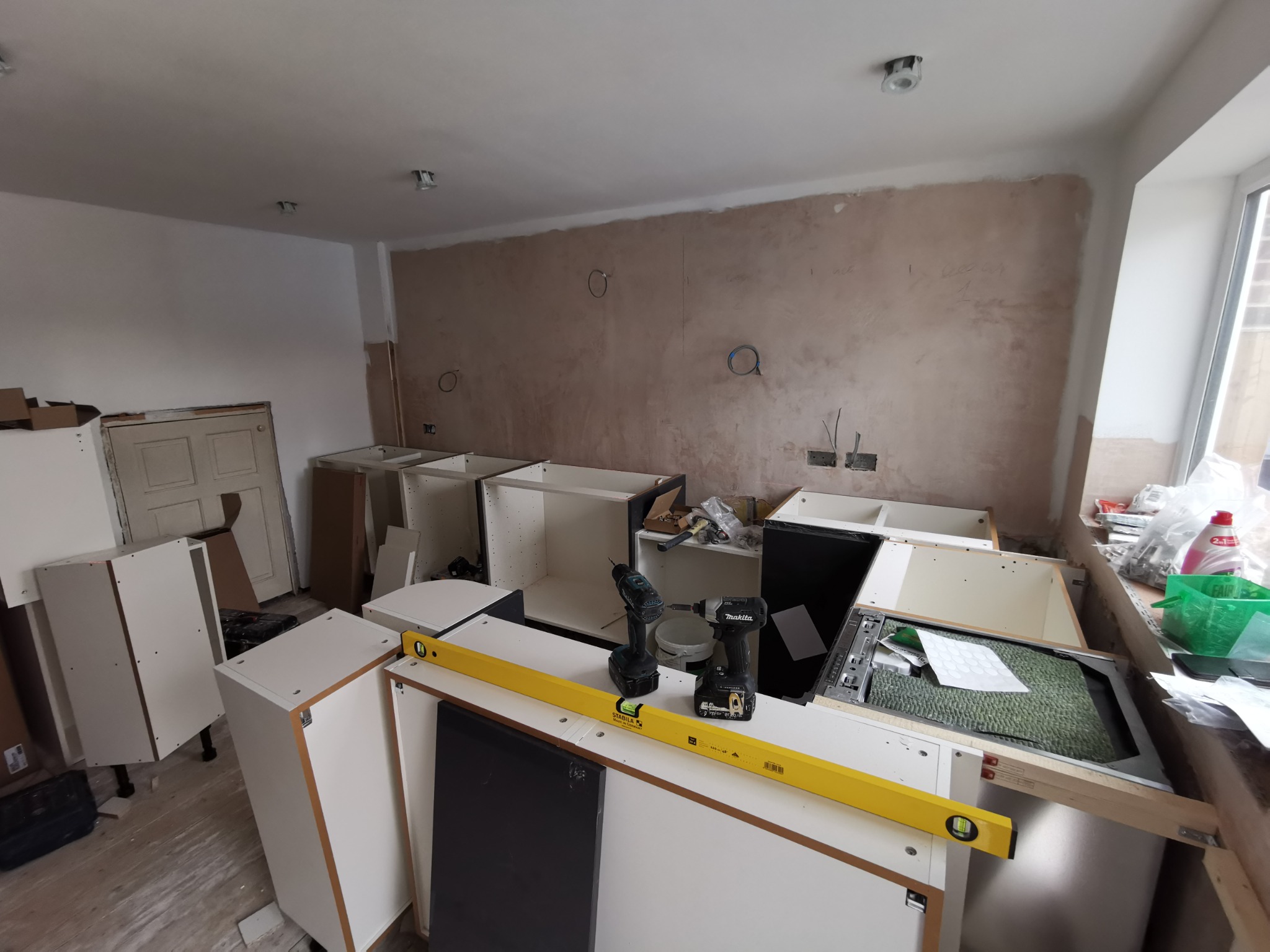 Wnętrze kuchni w trakcie montażu mebli: widoczne surowe ściany, częściowo złożone białe szafki, poziomica Stabila i dwa wkrętaki akumulatorowe Makita na blacie.