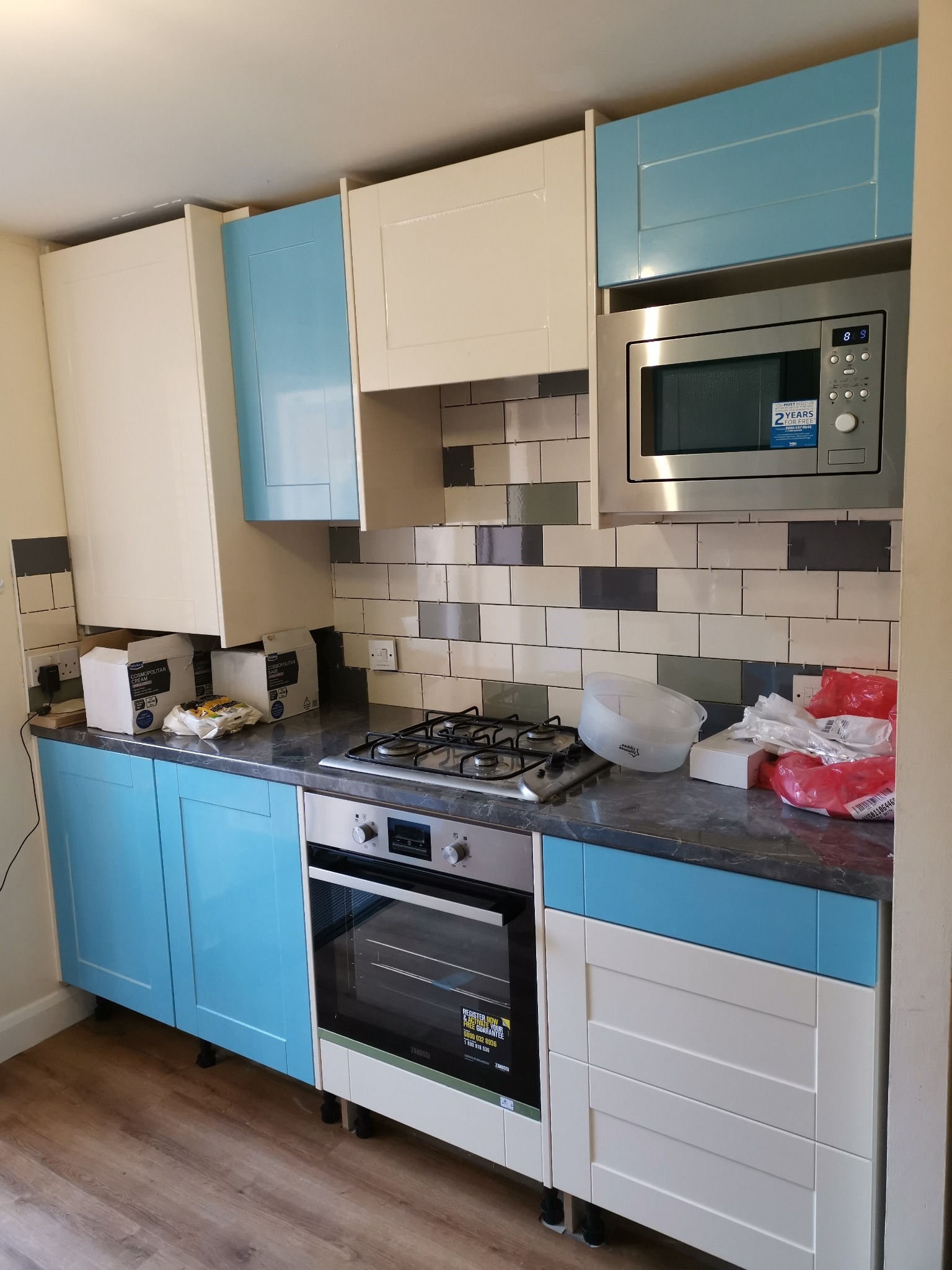 Nowoczesna kuchnia z białymi i błękitnymi szafkami, srebrnym mikrofalówką i piekarnikiem, oraz szarym blatem. Na blacie widoczne akcesoria i opakowania po montażu. Ściana wyłożona płytkami...