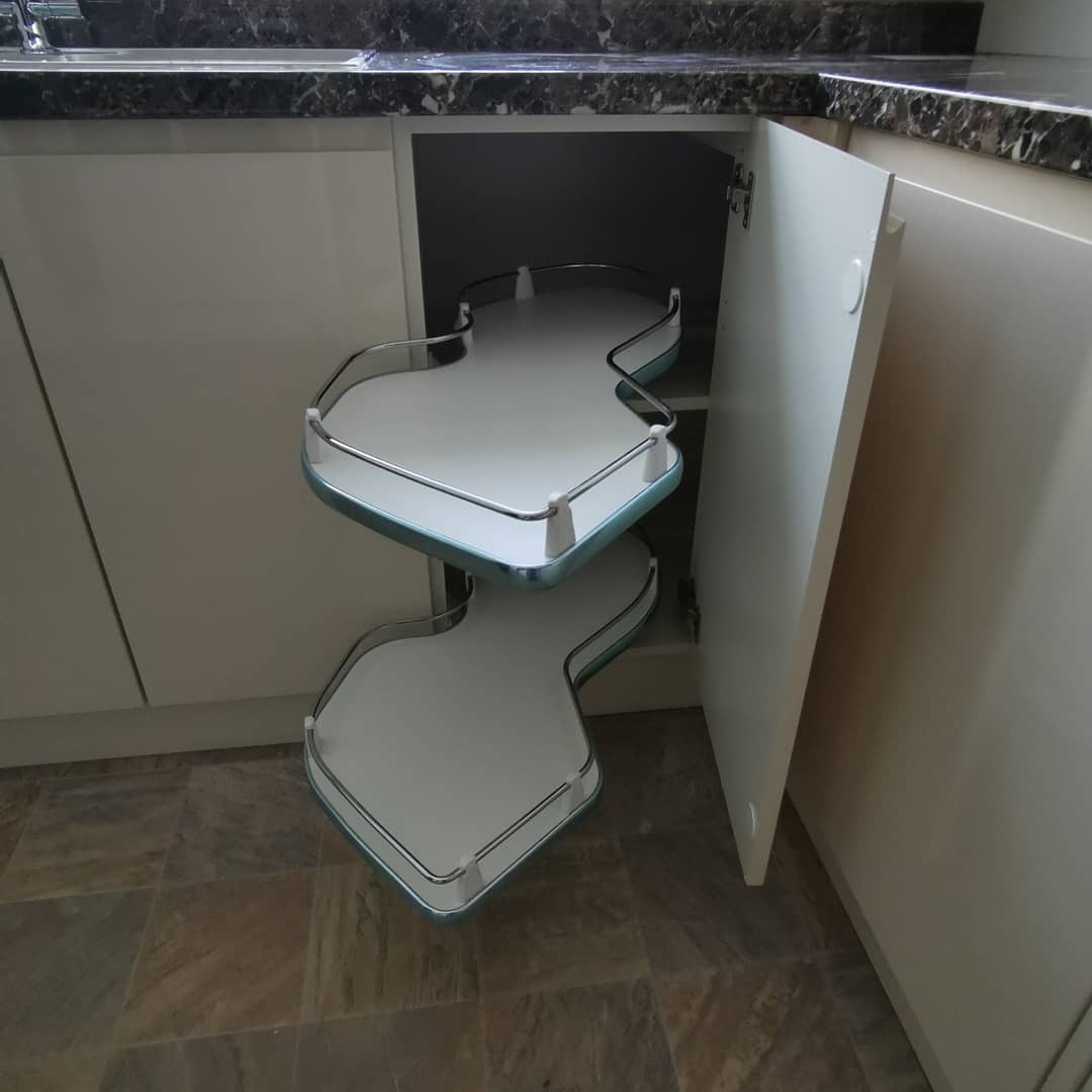 Wysuwana półka narożna w szafce kuchennej z otwartymi drzwiczkami, widoczne dwie białe półki z chromowanymi relingami i jasnoniebieskim obramowaniem.