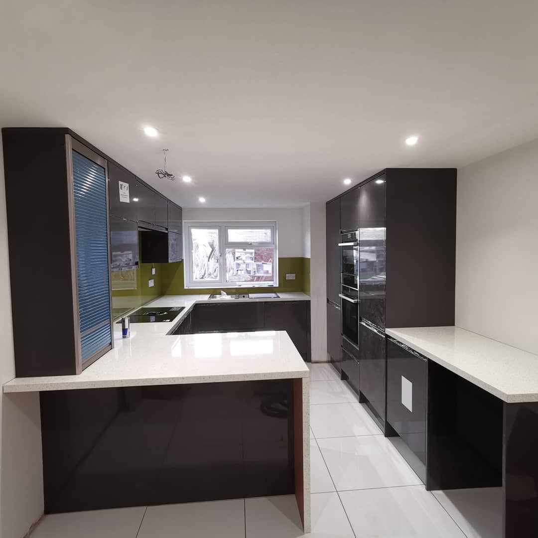 Nowoczesna kuchnia z ciemnymi, lakierowanymi frontami, białym blatem z konglomeratu i roletą w szafce narożnej, oświetlona punktowymi lampami sufitowymi.