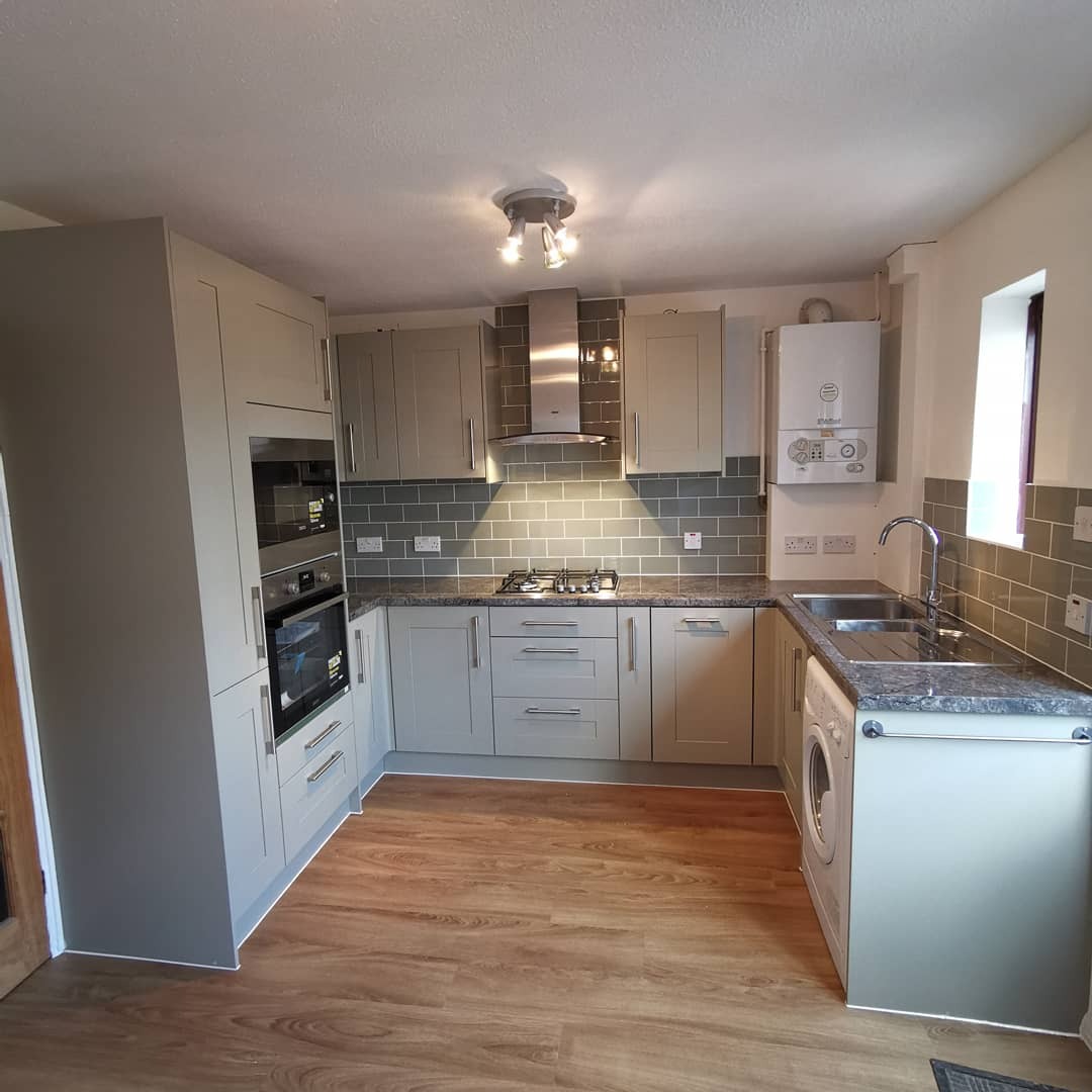 Jasnoszara kuchnia z drewnianą podłogą, szarymi płytkami nad blatem, z zamontowanym piecem gazowym na ścianie i pralką pod blatem.