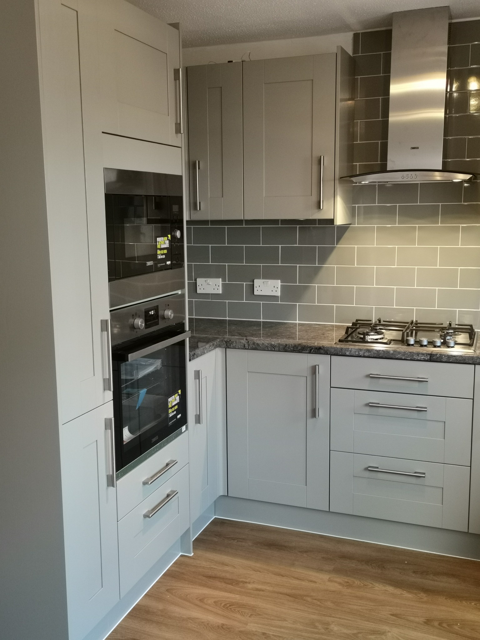 Szare szafki kuchenne z wbudowanym piekarnikiem i płytą gazową, szare płytki na ścianie, drewnopodobna podłoga, nowoczesny design.