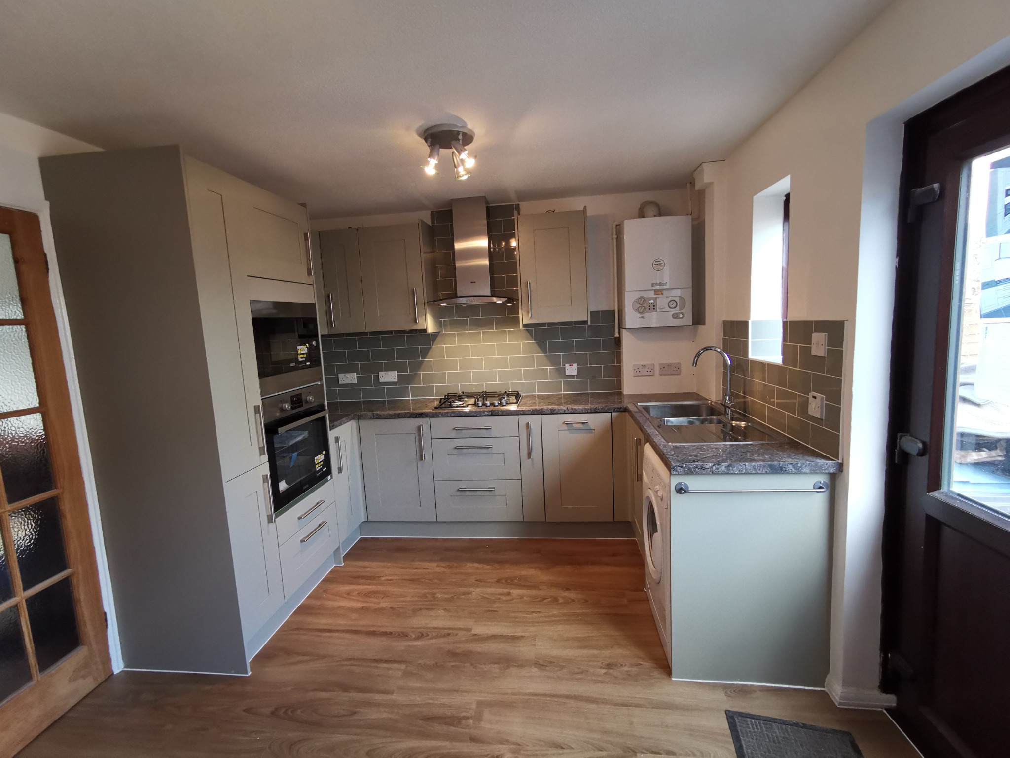 Ustawna kuchnia w odcieniach szarości z drewnopodobną podłogą, widoczny piec gazowy na ścianie oraz pralka w zabudowie.