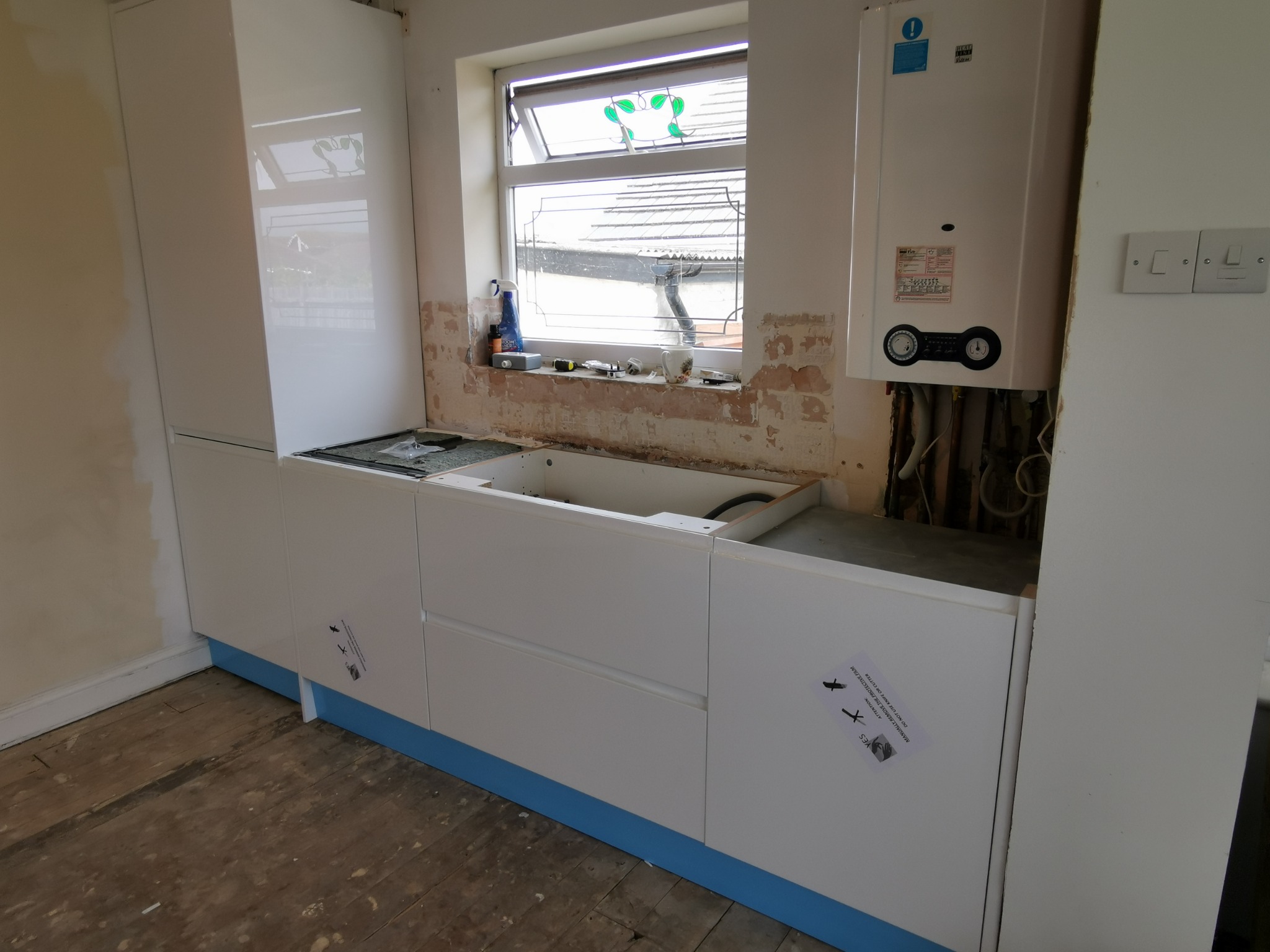 Montaż białych szafek kuchennych z szufladami w Braniewie, widoczne niedokończone ściany i piec gazowy nad blatem, na frontach naklejki ostrzegawcze o zdejmowaniu folii ochronnej.