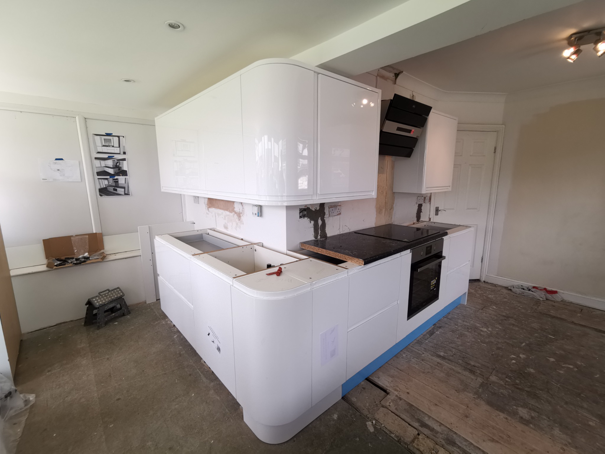 Montaż nowoczesnej, białej kuchni z zaokrąglonymi szafkami, widoczne niedokończone podłogi i przygotowane otwory na zlew, okap kuchenny już zamontowany.
