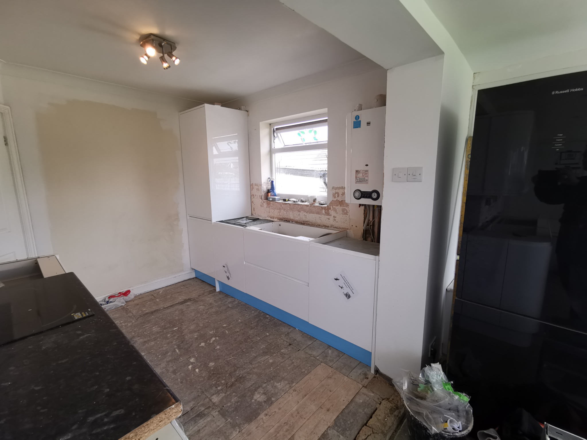 Wnętrze kuchni w trakcie remontu: białe szafki kuchenne z niebieskimi cokołami, surowe ściany, odsłonięta instalacja pod oknem, widoczny piec gazowy i czarna lodówka. Podłoga z desek w trakcie prac.