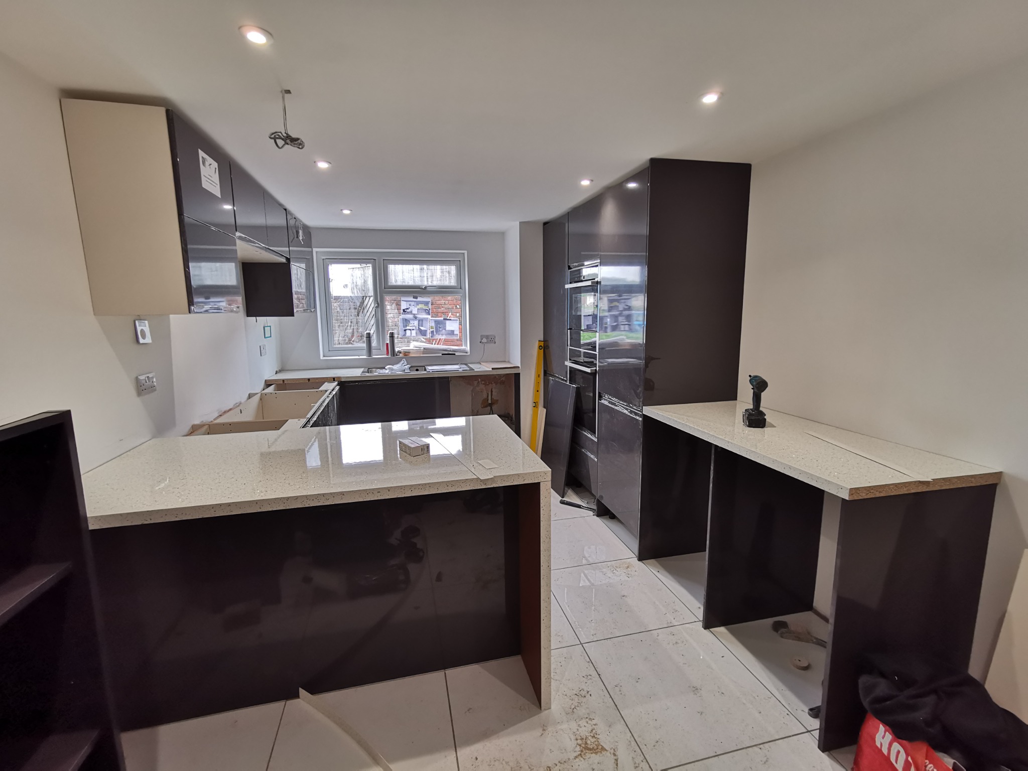 Wnętrze kuchni w trakcie montażu: ciemnobrązowe szafki z połyskiem, jasny blat z drobnym wzorem, widoczne narzędzia i elementy instalacji, okno z widokiem na budynek z cegły.