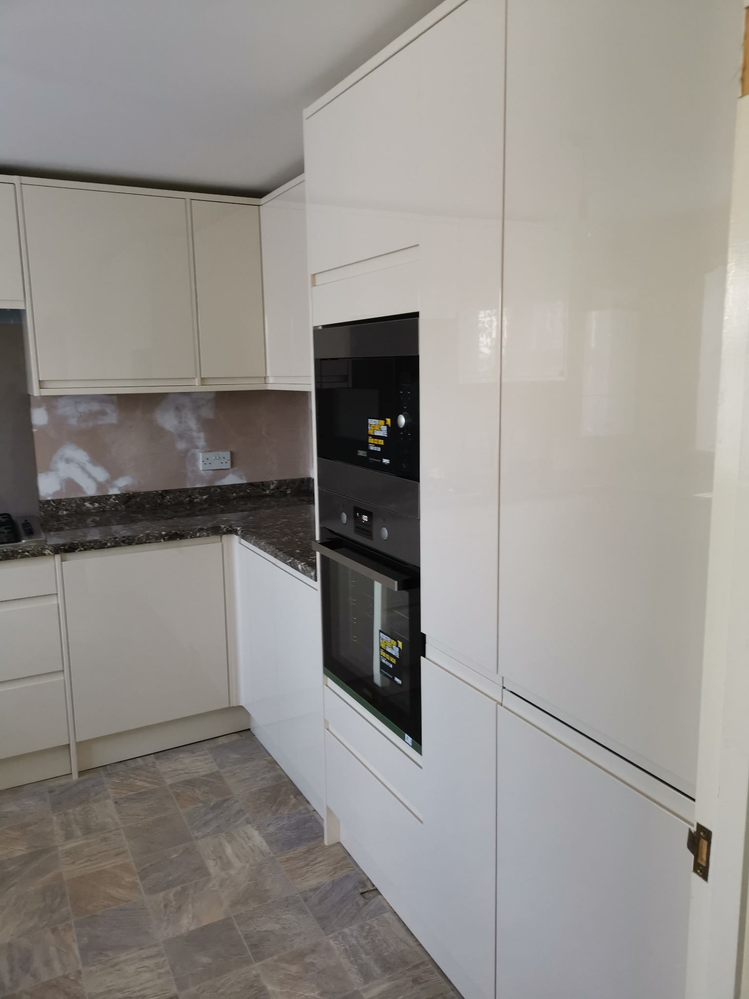 Biała, lakierowana zabudowa kuchenna z wbudowanym piekarnikiem i płytą grzewczą; ściana częściowo wykończona gładzią, widoczne gniazdko elektryczne; na podłodze płytki imitujące kamień.