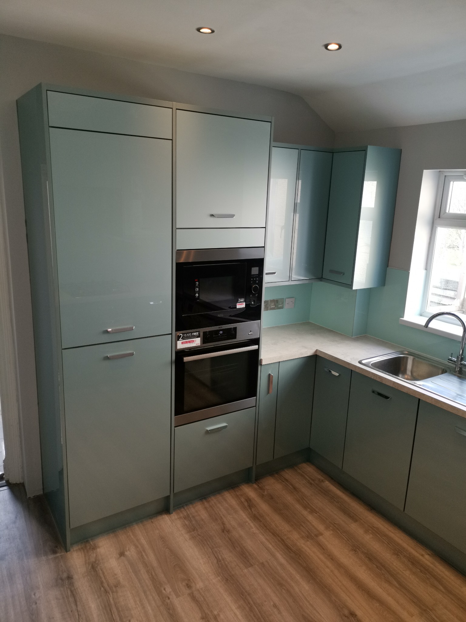 Nowoczesna kuchnia w odcieniach pastelowej zieleni z zabudowanymi sprzętami AGD, wykończona panelami podłogowymi o wzorze drewna.