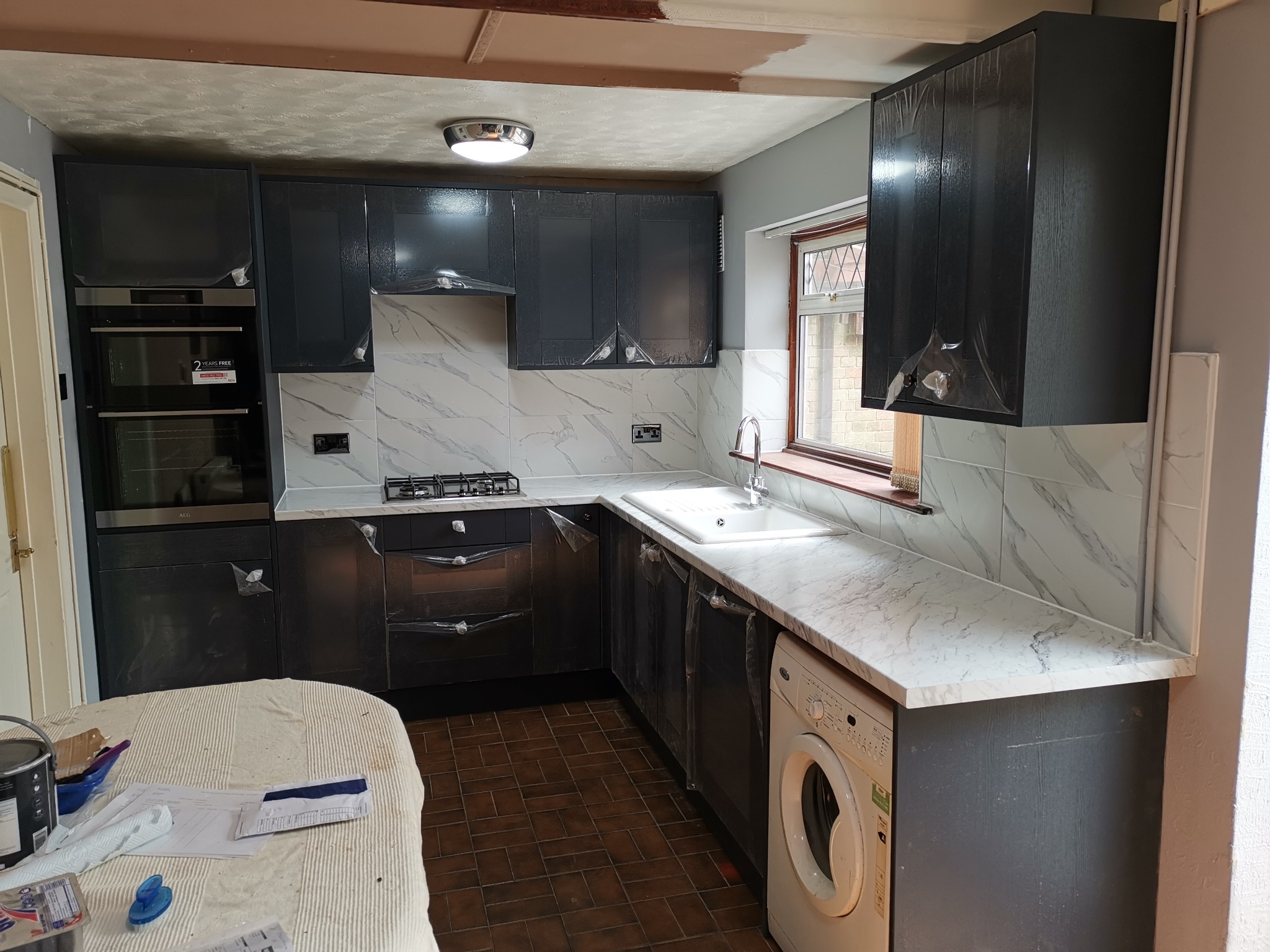 Ciemne szafki kuchenne z marmurowym blatem, nadal zabezpieczone folią ochronną po montażu, widoczna pralka pod blatem i stół z narzędziami malarskimi, sugerujące prace wykończeniowe.