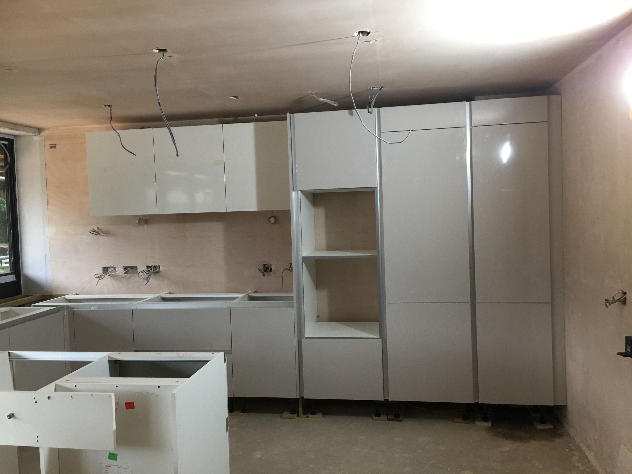 Montaż białych szafek kuchennych w surowym stanie, widoczne wystające kable ze ściany i sufitu, brak wykończenia ścian.