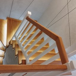 Usługi Stolarskie Paweł Żegociński - Widok z góry na drewniane schody z podświetleniem stopni i białą balustradą z drewnianą poręczą, na ścianie widoczny geometryczny wzór z czarnymi liniami.
