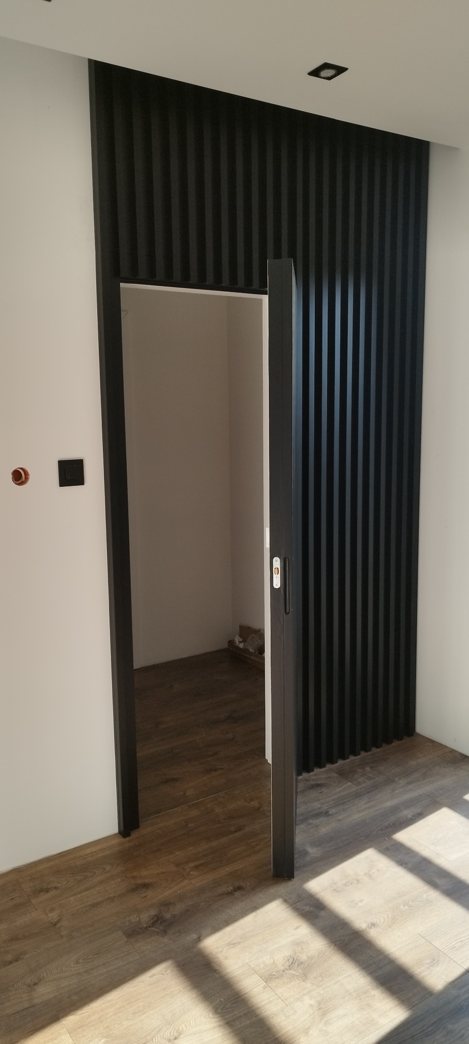 Otwarta, przesuwna, czarna drzwi ukryta z zamkiem, wkomponowana w ścianę z czarnych, pionowych lameli. Podłoga z paneli imitujących drewno. Widoczny fragment instalacji elektrycznej.