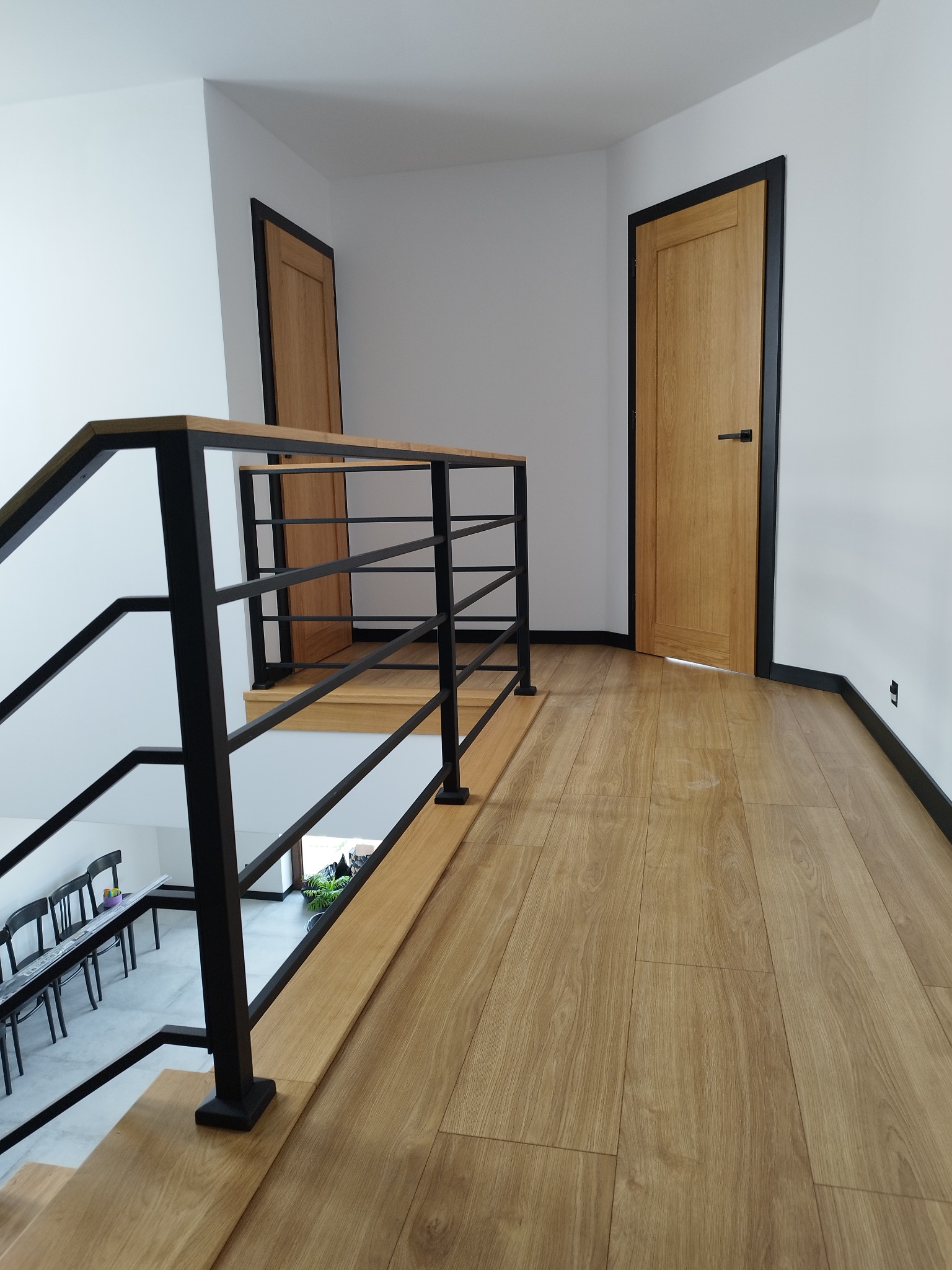 Wnętrze domu z drewnianymi schodami z czarną metalową balustradą i dwoma drewnianymi drzwiami z czarnymi ościeżnicami na białych ścianach, widok z góry schodów na korytarz.
