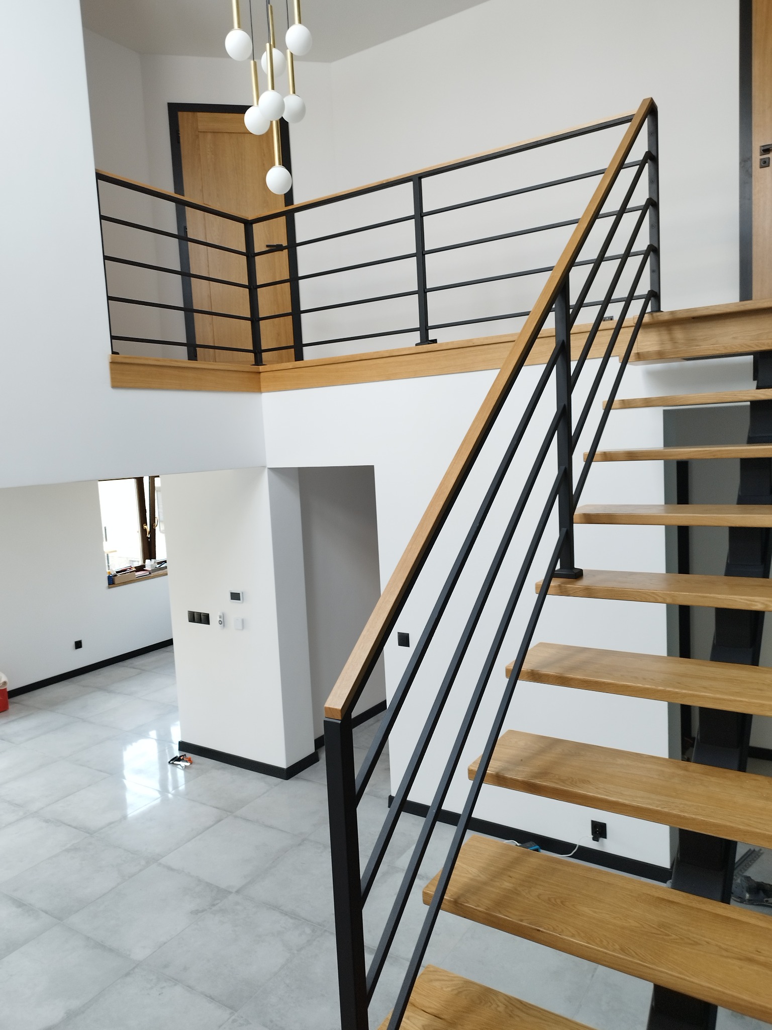 Nowoczesne schody z dębowymi stopniami na metalowej konstrukcji i czarną balustradą, widok z dołu, w tle antresola z identyczną balustradą, białe ściany i szara podłoga.