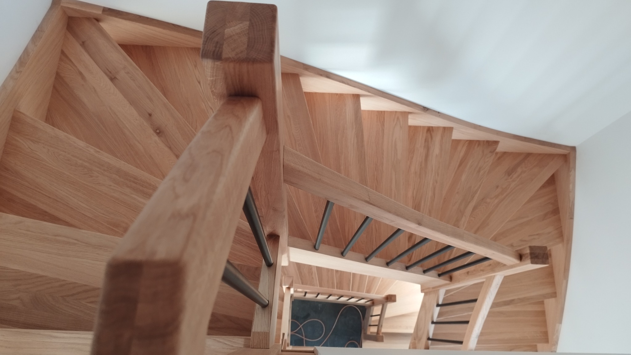Spiralne schody z jasnego drewna z metalowymi poręczami, widok z góry, ukazujący zakrzywiony bieg stopni i minimalistyczny design.
