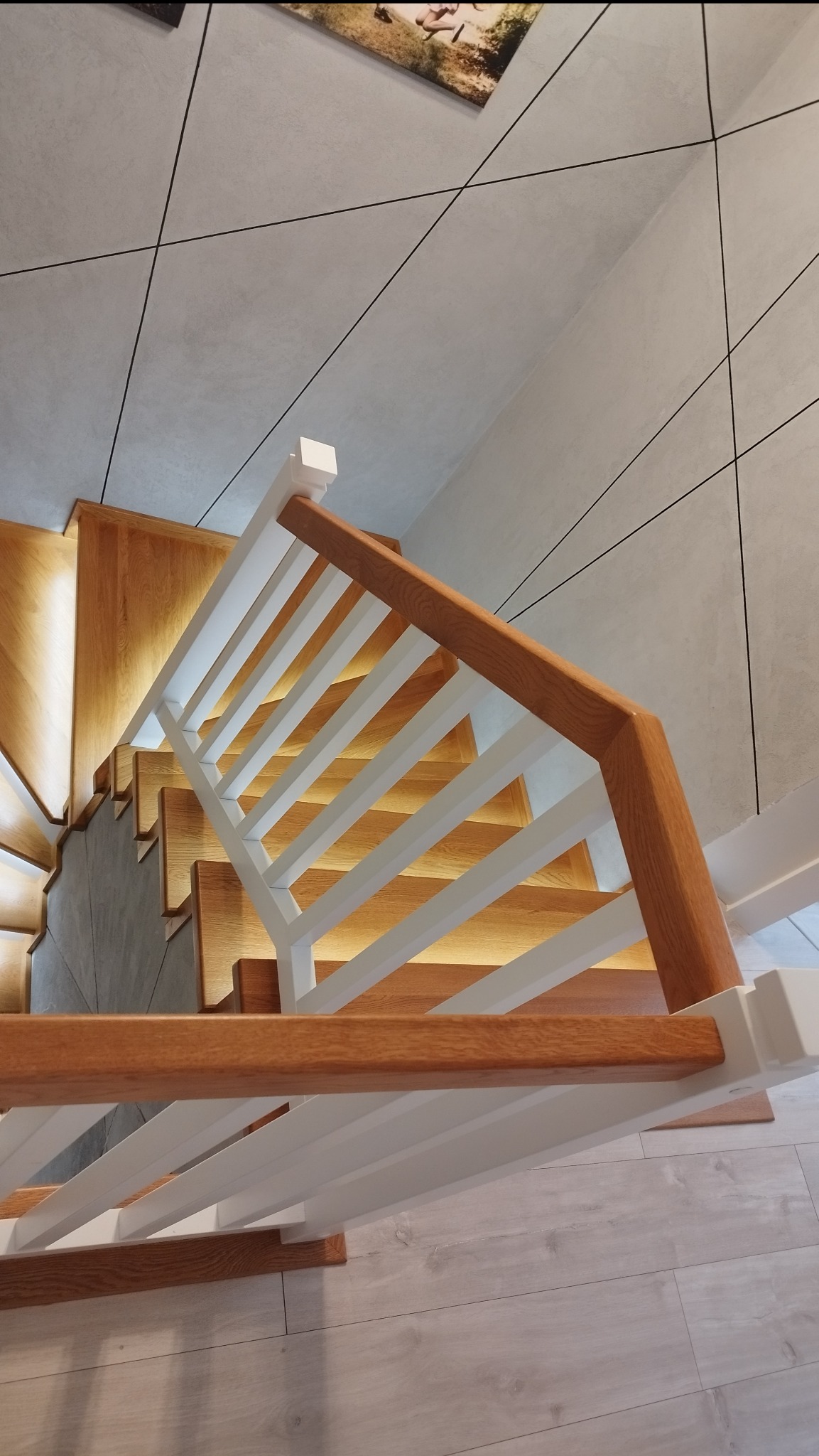 Widok z góry na drewniane schody z podświetleniem stopni i białą balustradą z drewnianą poręczą, na ścianie widoczny geometryczny wzór z czarnymi liniami.