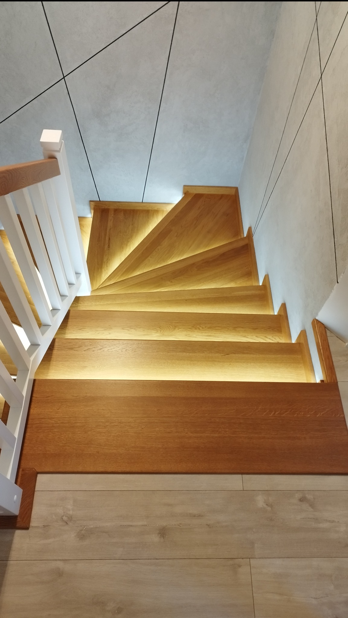 Widok z góry na dębowe schody z podświetleniem LED pod stopniami i białą balustradą, nowoczesny design z geometrycznym wzorem na suficie i ścianie.