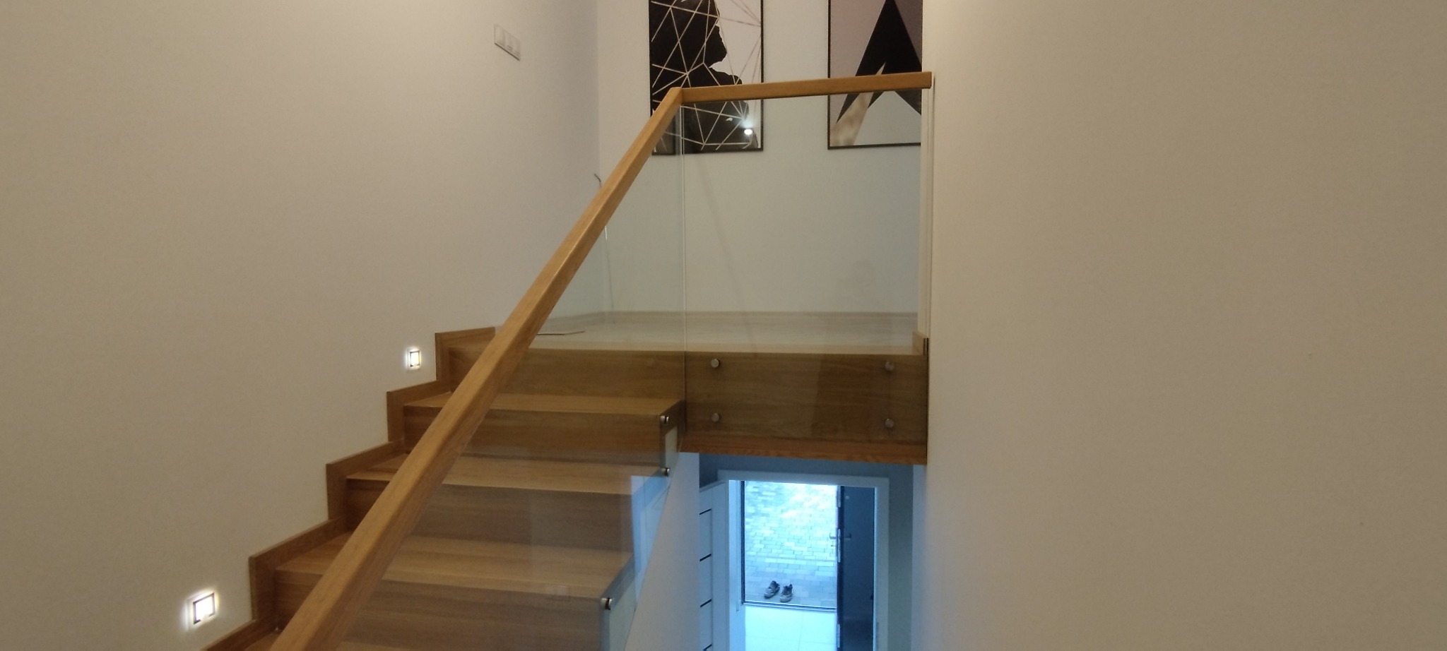Schody dywanowe z dębowymi stopniami i szklaną balustradą, oświetlenie punktowe w ścianie, minimalistyczne grafiki na ścianie w tle.