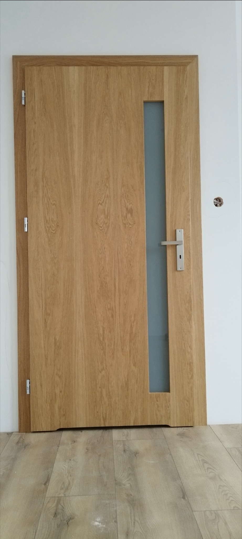 Zamknięte, fornirowane drzwi wewnętrzne z wąskim, pionowym przeszkleniem, zamontowane w białej ścianie. Widoczna klamka i zawiasy. Podłoga wyłożona panelami imitującymi drewno.