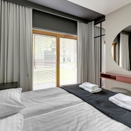 Projekt 5R - Nowoczesna sypialnia z łóżkiem dwuosobowym, pościel w paski, czerwone wezgłowie, okrągłe lustro nad toaletką z szufladą, szare zasłony i rolety w oknie.